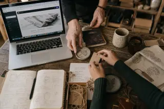 Artisan hands arranging products beside a laptop showing a website editor, in a softly lit, authentic workshop