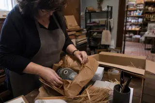 Shop owner's hands carefully wrapping a unique handmade item in a warmly lit back room, shelves of local products visible in the background