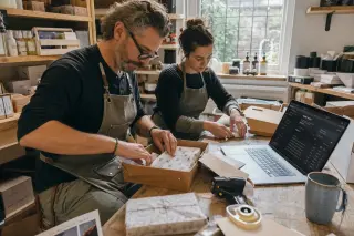 Shopkeeper and assistant packing orders in a cluttered store room, focused on their work, with a laptop and products around them under gentle natural light.